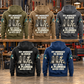 Five complete back views of the Plumber Never Ends Vintage Hoodie in Military Green, Kaki, Dark Gray, Black and Dark Blue, arranged in a plumbing workshop with pipe fittings and tools behind the garments.