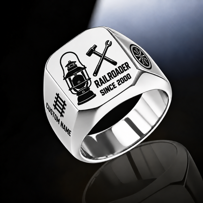 Silver Railroader Split Emblem ring with an asymmetric railway lantern and crossed railway-tool composition, with the name on the left shoulder below a track motif and a rail-wheel emblem on the right shoulder, with illustrative CUSTOM NAME and SINCE 2000 artwork