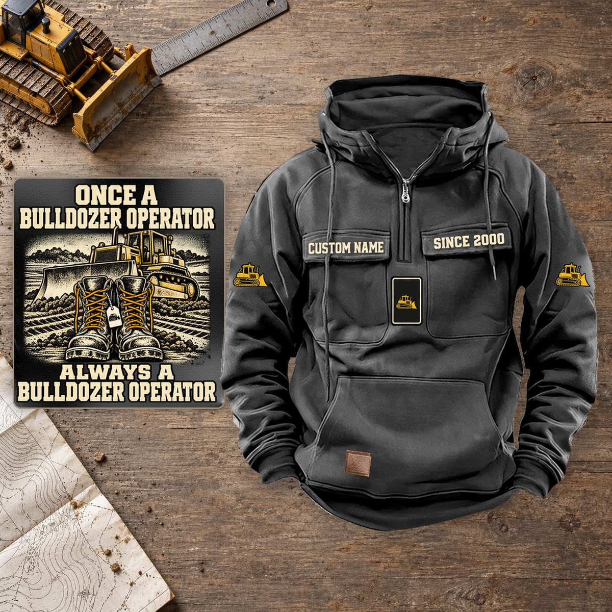 Bulldozer Operator Once and Always Vintage Hoodie in Dark Gray, front view with sample name and year panels, plus a close-up of the boots-and-bulldozer back artwork on a workshop table.