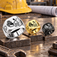 Silver-dominant Bulldozer Operator Split Emblem signet rings in Silver, Gold and Black on stepped steel blocks on a stationary workshop tabletop; tracked bulldozer and crossed-tool artwork with sample CUSTOM NAME and SINCE 2000.