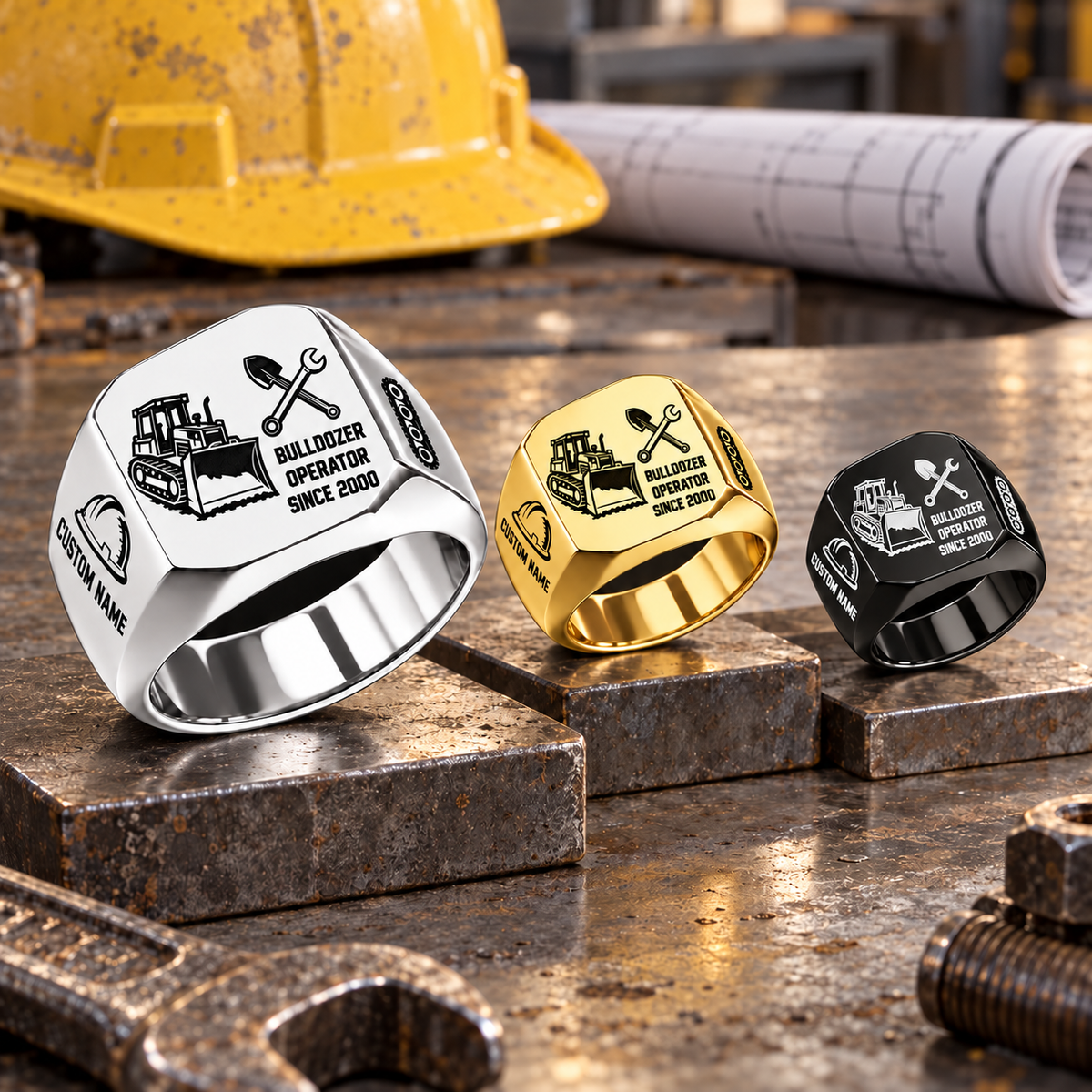 Silver-dominant Bulldozer Operator Split Emblem signet rings in Silver, Gold and Black on stepped steel blocks on a stationary workshop tabletop; tracked bulldozer and crossed-tool artwork with sample CUSTOM NAME and SINCE 2000.