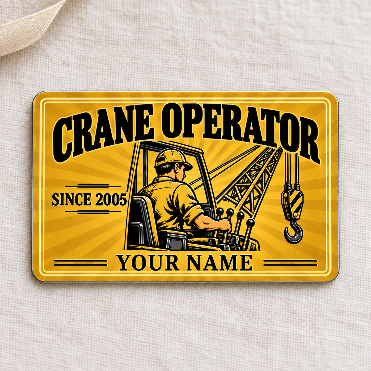 Front of the Crane Operator Golden Milestone aluminum wallet card on cream linen, showing an operator at crane controls, lattice boom, hook, and illustrative SINCE 2005 and YOUR NAME text.