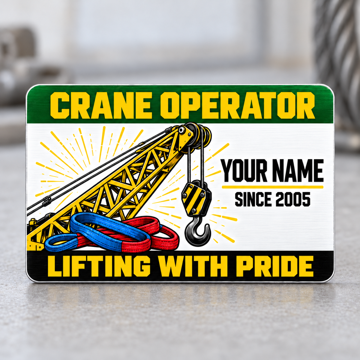 Front of the Crane Operator Green Band aluminum wallet card upright on a pale gray tabletop, with a yellow crane boom, hanging hook, blue and red rigging slings, green header and black Lifting with Pride footer.