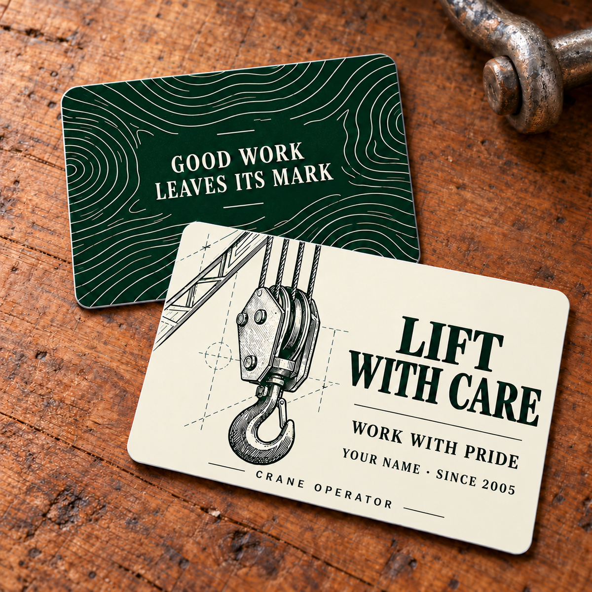 Front and reverse of the Crane Operator Lift with Care printed aluminum wallet card on a workbench: ivory crane-hook technical artwork with Lift with Care and Work with Pride, and a dark green woodgrain reverse reading Good Work Leaves Its Mark. Styling props are not included.