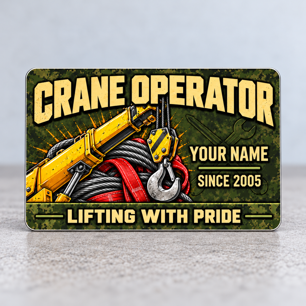 Front of the Crane Operator Rugged Field aluminum wallet card standing upright on a gray tabletop, with olive pixel camouflage, tan lettering, yellow crane boom and hook, gray wire rope, red lifting sling, and illustrative name and year.