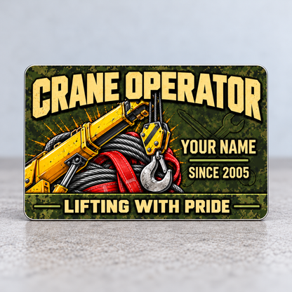 Front of the Crane Operator Rugged Field aluminum wallet card standing upright on a gray tabletop, with olive pixel camouflage, tan lettering, yellow crane boom and hook, gray wire rope, red lifting sling, and illustrative name and year.