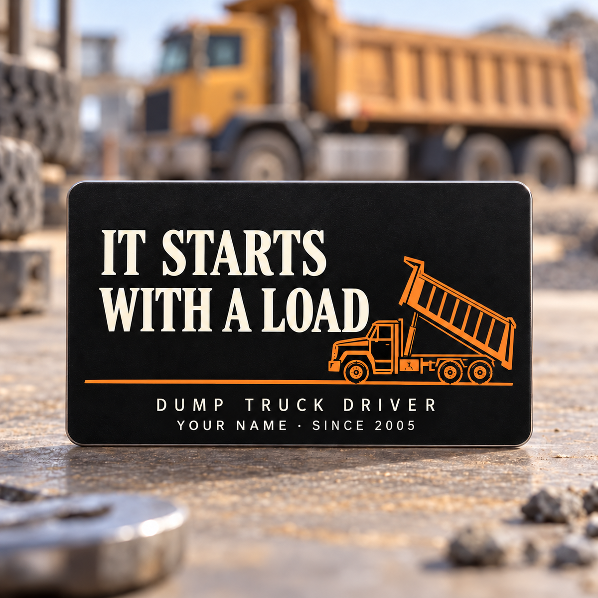 Black front of the Dump Truck Driver First Load aluminum wallet card, showing It Starts with a Load in ivory and an orange rigid dump truck with its tipping body raised.