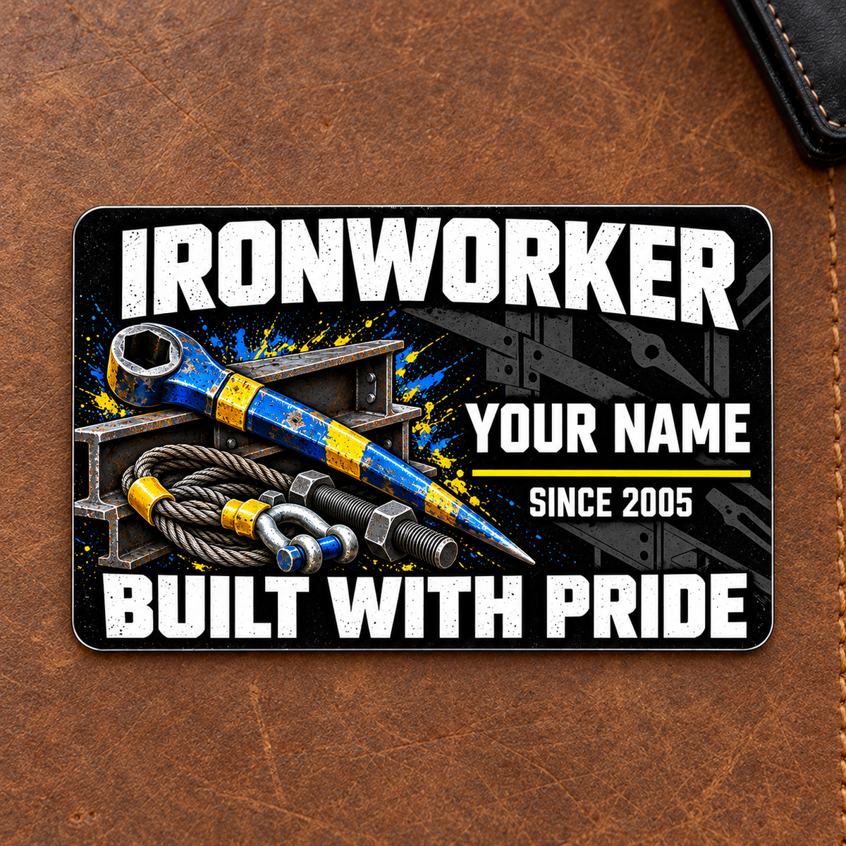 Ironworker Midnight Pride aluminum wallet card front on a brown leather mat, with distressed white lettering and blue-yellow spud wrench, I-beams, bolts and lifting sling artwork.