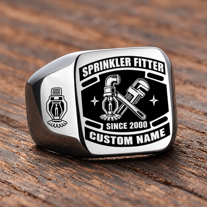 Silver-finish Sprinkler Fitter Black Face Badge ring with fire-sprinkler head, pipe and wrench artwork, shoulder sprinkler emblem and illustrated CUSTOM NAME and SINCE 2000 text