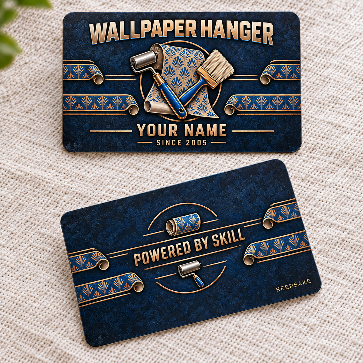 Front and reverse views of one Wallpaper Hanger Craft Camo aluminum wallet card on cream woven fabric, showing navy textured camouflage, copper-toned lettering, blue accents, wallpaper rolls, seam roller, smoothing brush and curled patterned-paper borders.