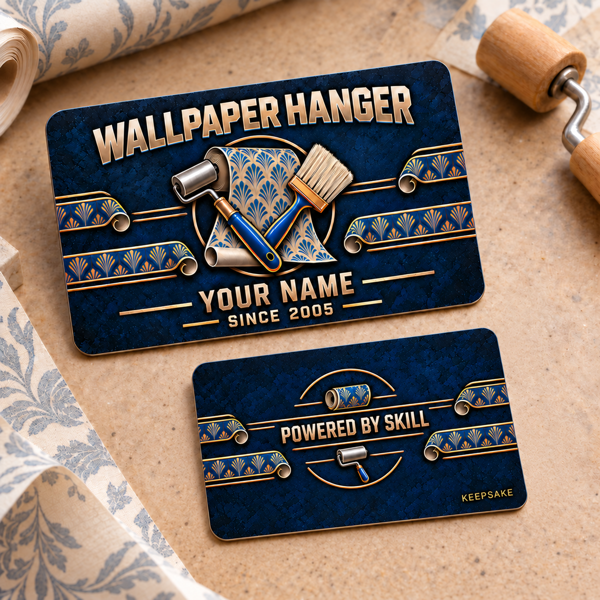 Front and reverse of one Wallpaper Hanger Craft Camo aluminum wallet card on a wallpaper preparation tabletop, with navy textured camo, copper and blue wallpaper rolls, seam roller, smoothing brush and patterned-paper borders. Illustrative YOUR NAME text appears on the front; the reverse reads Powered by Skill and Keepsake.