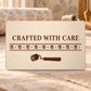 Ivory reverse of the Wallpaper Hanger Every Seam aluminum wallet card, reading Crafted with Care above a repeating leaf-and-diamond wallpaper-pattern band and brown seam-roller silhouette.
