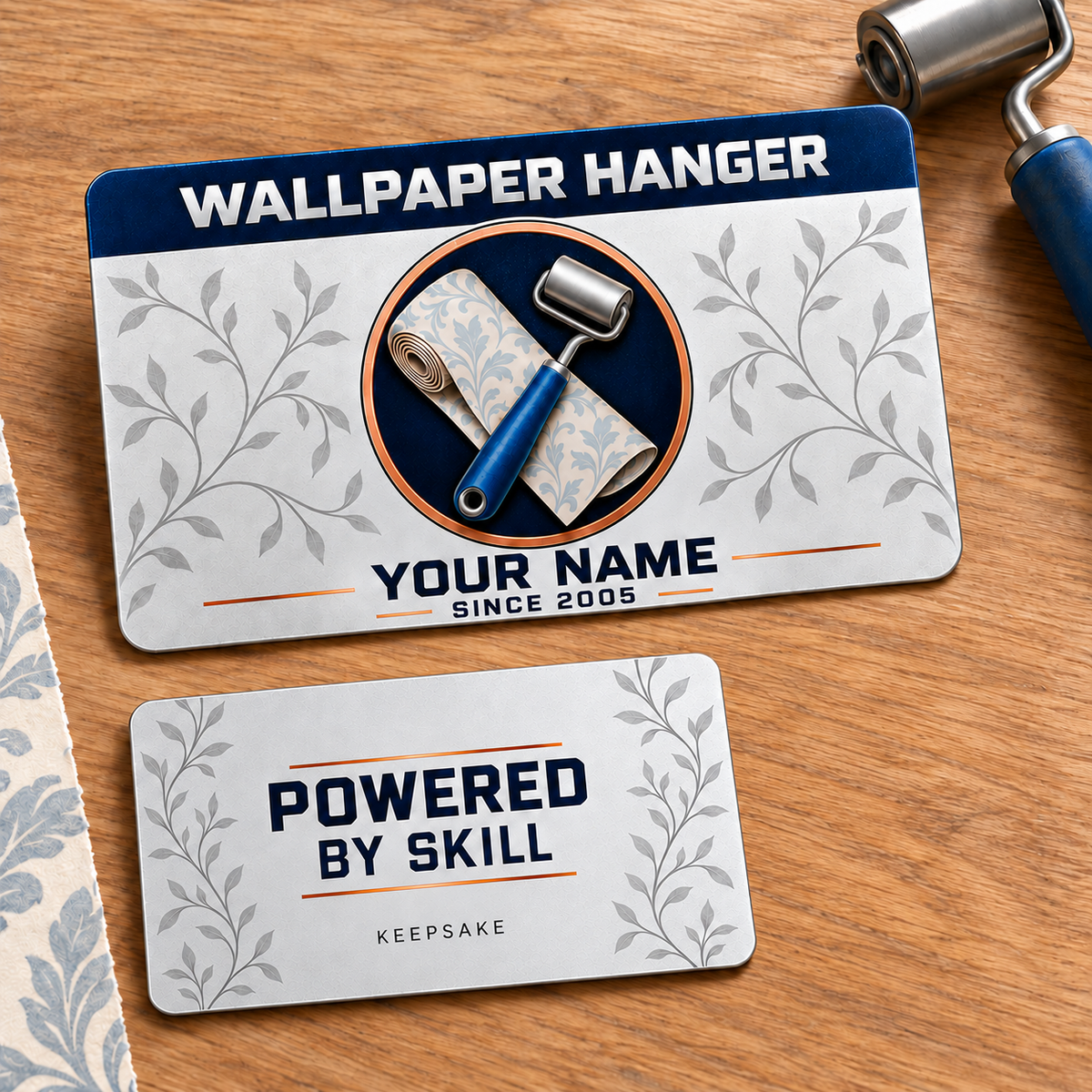 Front and reverse views of one Wallpaper Hanger Everyday Craft aluminum wallet card on an oak wallpaper tabletop. Silver leaf-vine artwork surrounds a wallpaper roll and seam roller medallion; illustrative YOUR NAME text is shown on the front.