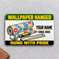Front of the Wallpaper Hanger Green Band aluminum wallet card on light gray fabric, with a floral wallpaper roll, blue and yellow seam roller, rule, and illustrative YOUR NAME and SINCE 2005 text.