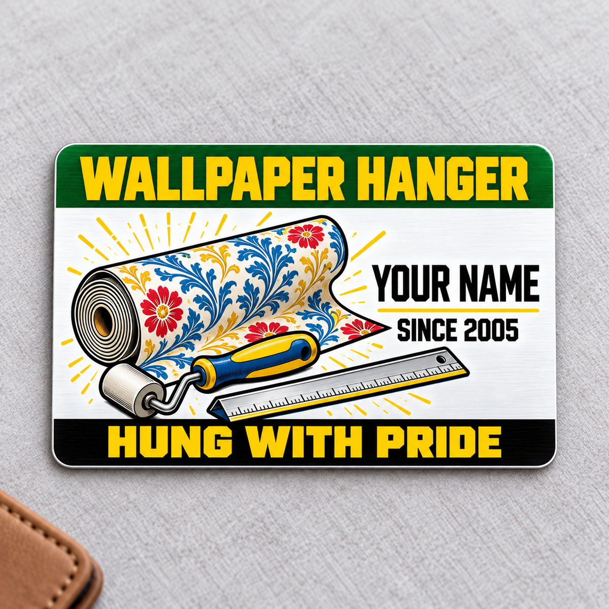 Front of the Wallpaper Hanger Green Band aluminum wallet card on light gray fabric, with a floral wallpaper roll, blue and yellow seam roller, rule, and illustrative YOUR NAME and SINCE 2005 text.