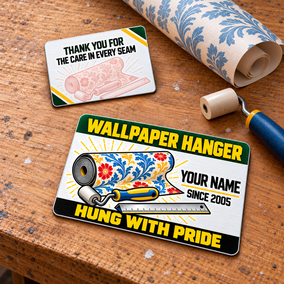 Front and reverse of one Wallpaper Hanger Green Band aluminum wallet card displayed diagonally on a wallpaper workbench; green and yellow header, floral wallpaper roll, seam roller and rule. Name and year are illustrative design text.