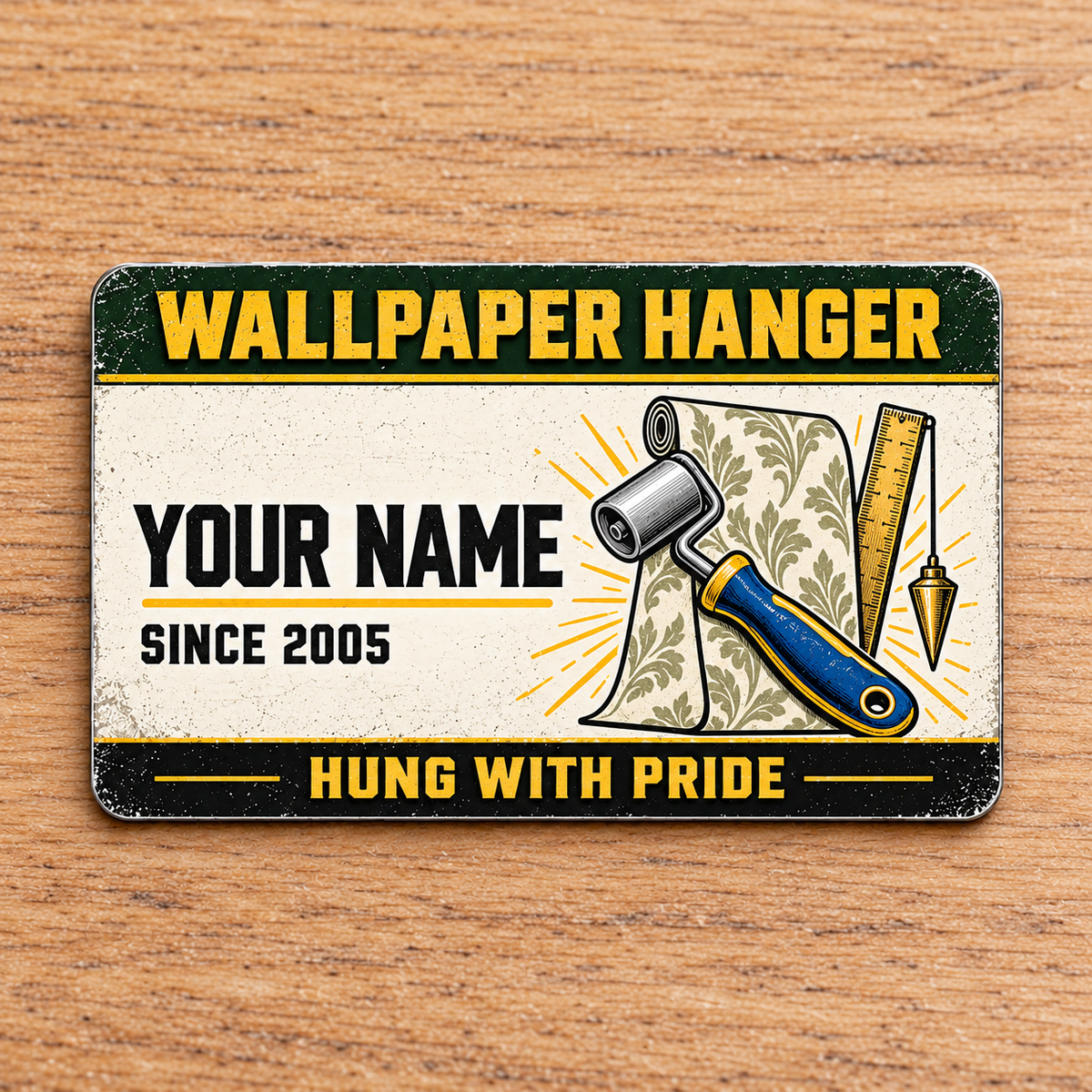 Front of the Wallpaper Hanger Heritage Band aluminum wallet card with green and gold bands, illustrative name and year, patterned wallpaper roll, seam roller, ruler and plumb bob.
