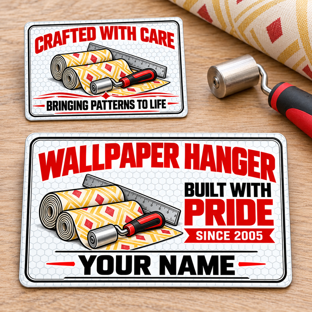 Offset front and reverse views of one Wallpaper Hanger Honeycomb Pride aluminum wallet card on a light oak wallpaper worktable. Pale honeycomb, red and black lettering, patterned yellow-red wallpaper rolls, a seam roller and measuring rule appear on both faces; YOUR NAME and SINCE 2005 are illustrative text.