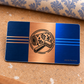 Overhead reverse view of the Wallpaper Hanger Midnight Pride aluminum wallet card, with fine brushed metallic navy and blue side panels, a copper central stripe, black wallpaper roll and seam roller emblem, straight decorative stripes and KEEPSAKE lettering.