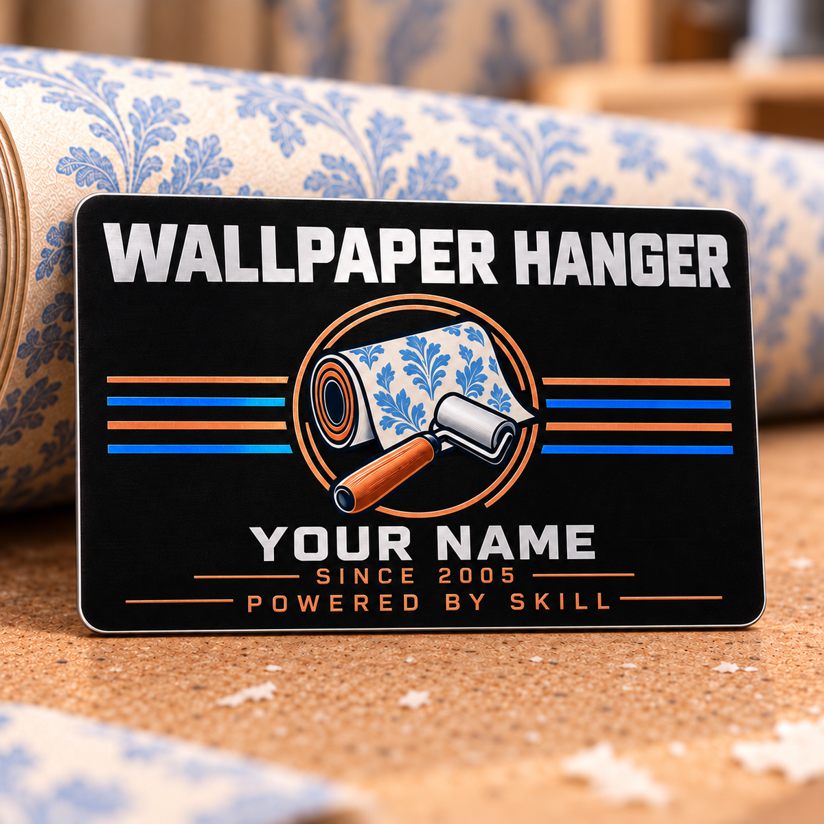 Wallpaper Hanger Midnight Pride aluminum wallet card standing upright on a wallpaper preparation table. The black front has silver Wallpaper Hanger and illustrative YOUR NAME lettering, a blue-patterned wallpaper roll and copper-handled seam roller, copper circular frame, straight copper and blue stripes, and Powered by Skill lettering.