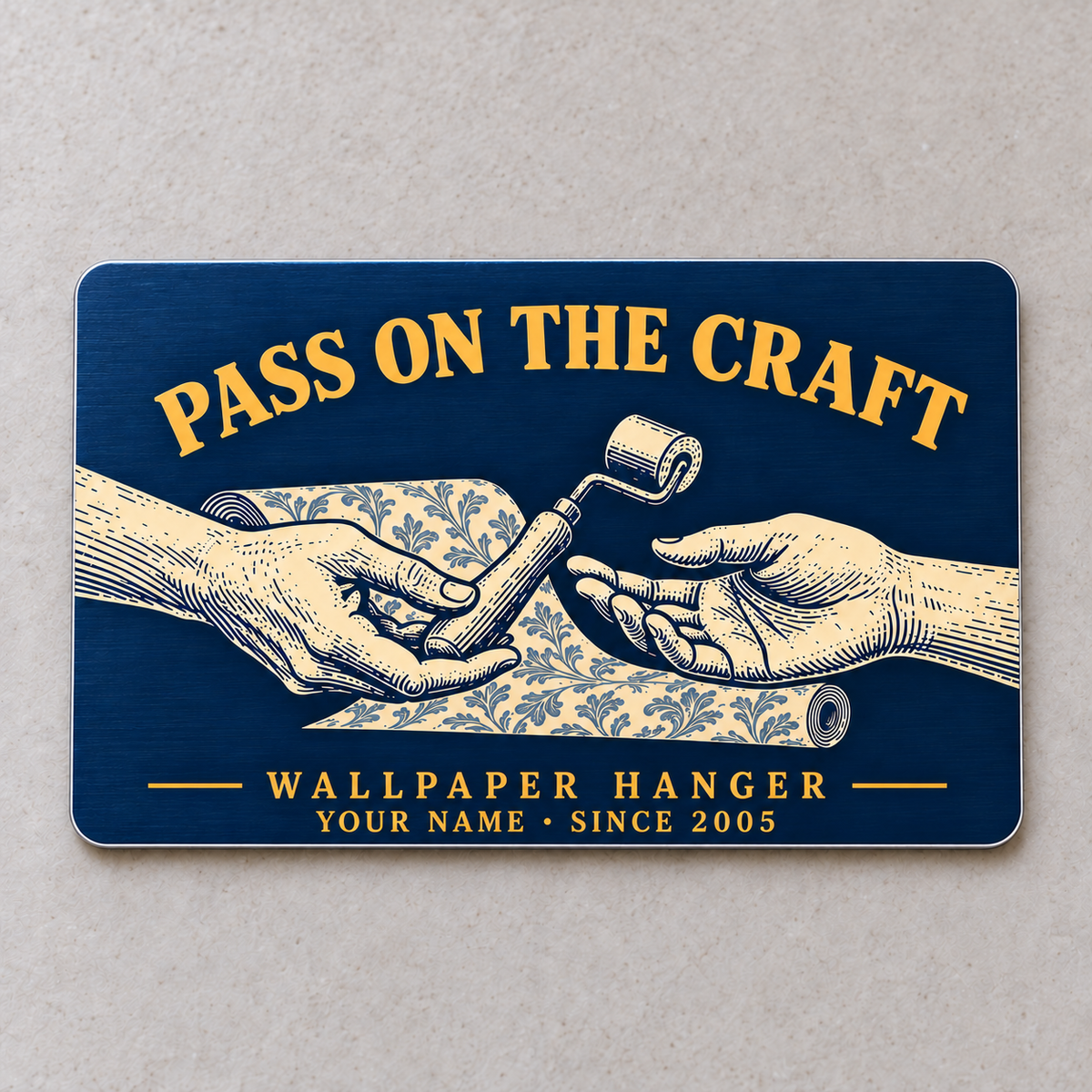 Navy front of the Wallpaper Hanger Pass on the Craft aluminum wallet card, showing two hands passing a seam roller over patterned wallpaper with gold lettering.