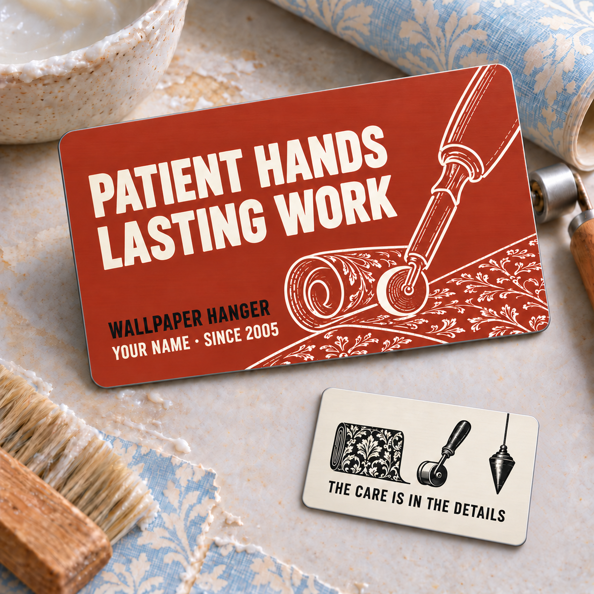 Wallpaper Hanger Patient Hands two-sided aluminum wallet card, with brick-red front and smaller ivory reverse arranged diagonally on a wallpaper worktable.