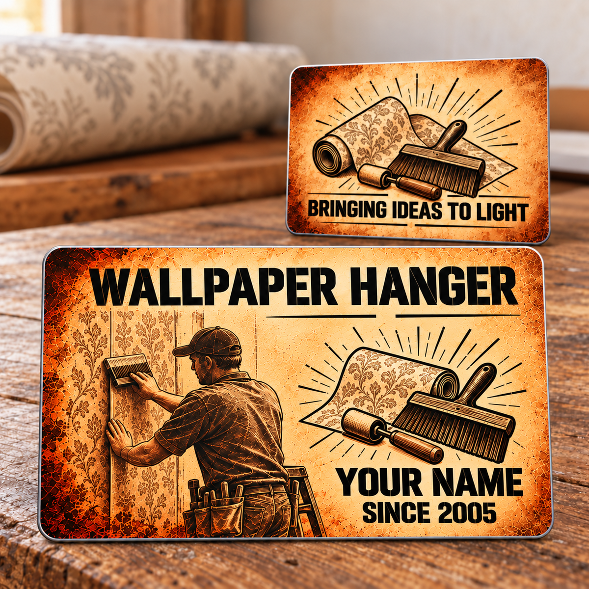 Two views of the Wallpaper Hanger Sepia Legacy aluminum wallet card on a timber workbench, with a sepia wallpaper worker, wallpaper roll, seam roller and smoothing brush.