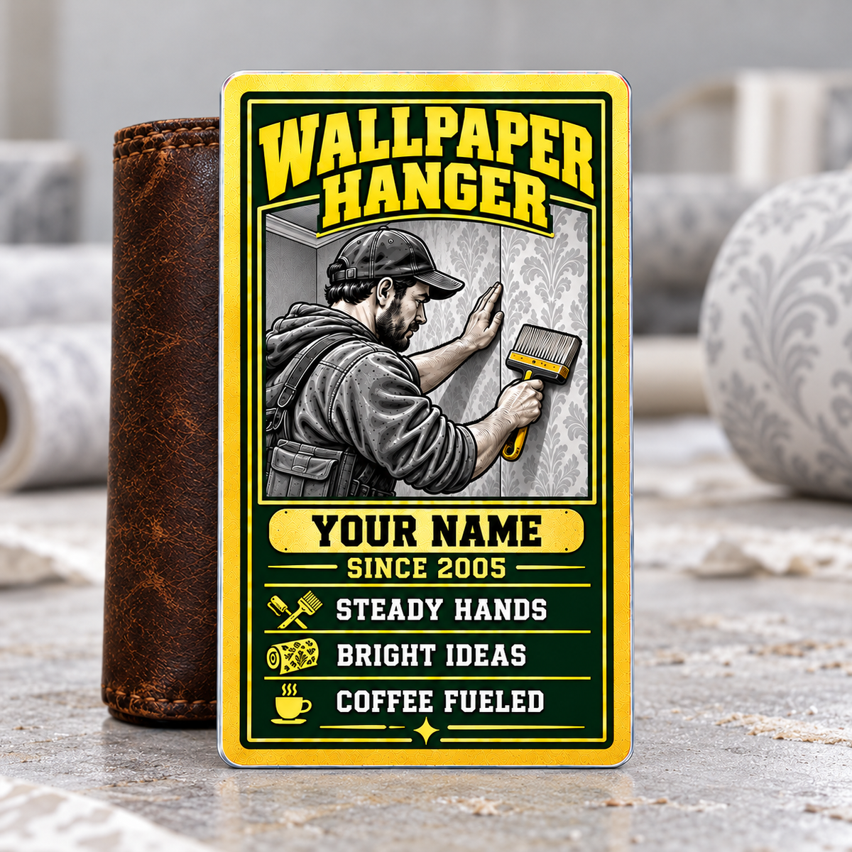 Wallpaper Hanger Trade Legend aluminum wallet card, portrait front with green-and-gold artwork, supported by a brown wallet on a wallpapering table