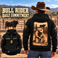 Black Bull Rider Daily Commitment hoodie on a cowboy at a corral, showing the sepia kneeling-rider back print and a smaller front view.