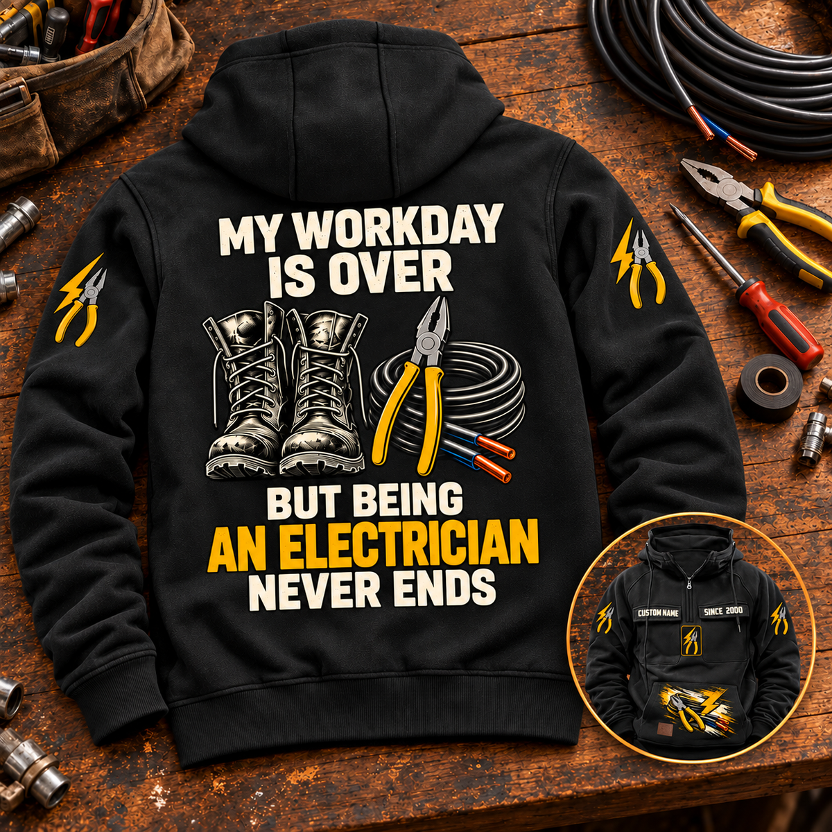 Black Electrician Never Ends Vintage Hoodie laid back-up on a dark timber workbench, with readable boots, pliers and cable artwork and a smaller front-view inset.
