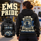 Black EMS Heritage Crest Vintage Hoodie worn by an adult woman outside an EMS station, showing the large cream-and-blue back crest and a smaller front-view inset with CUSTOM NAME and SINCE 2000.