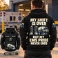 Black EMS Never Ends Vintage Hoodie worn from behind at a quiet ambulance station, with boots, ambulance and response bag artwork, readable EMS pride slogan, and a smaller complete front view.