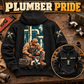 Black plumber hoodie on a workshop bench, showing the kneeling plumber and pipework back print with a smaller front-view inset.