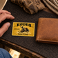 Bull Riding/Rodeo Golden Milestone aluminum wallet card on dark felt beside a tan wallet and a relaxed adult hand