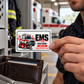 Adult male responder holding the Honeycomb Pride EMS card by its edge at a station locker, above an open brown wallet.