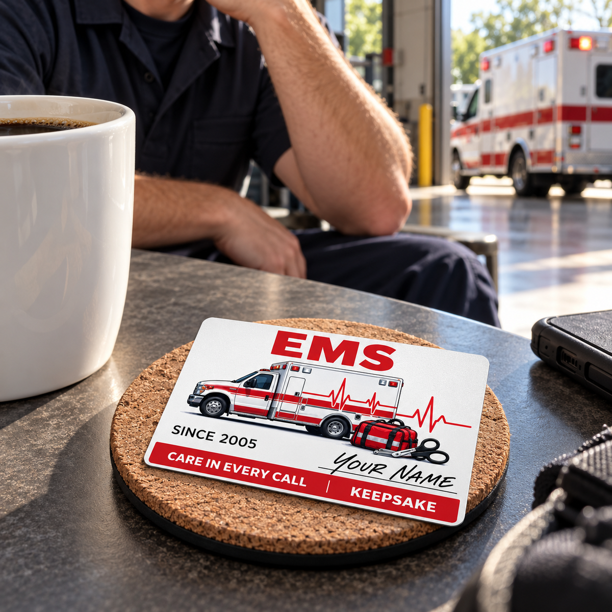 EMS Signature Classic card on a cork coaster beside coffee, with an adult responder seated behind at the station.