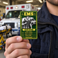 EMS Trade Legend portrait wallet card held by an adult worker near a parked ambulance.