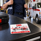 Signature Modern EMS keepsake card lying flat on a dark tool cabinet while an adult responder puts on a jacket nearby.