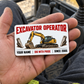 The printed front of the Excavator Operator Minimal Tool Grid aluminum wallet card shown in an occupational lifestyle setting with an adult worker.