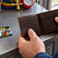 HVAC Heritage Band aluminum wallet card lying flat beside an open brown bi-fold wallet held by an adult technician at an HVAC workshop shelf.