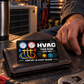 HVAC Midnight Pride aluminum wallet card resting on a closed brown leather wallet beside an adult worker's hand in a warmly lit HVAC workshop.