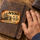 HVAC Sepia Legacy aluminum keepsake wallet card lying on a worn work journal beside an adult hand and brown leather wallet on a wooden bench.