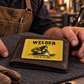 Welder Golden Milestone wallet card resting on a closed leather wallet beside an adult welder's hands in a workshop