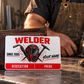 Welder Signature Modern card leaning against a wallet on a workshop bench