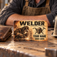 Welder Sepia Legacy aluminum wallet card beside a worn wallet and welding hood in warm workshop light